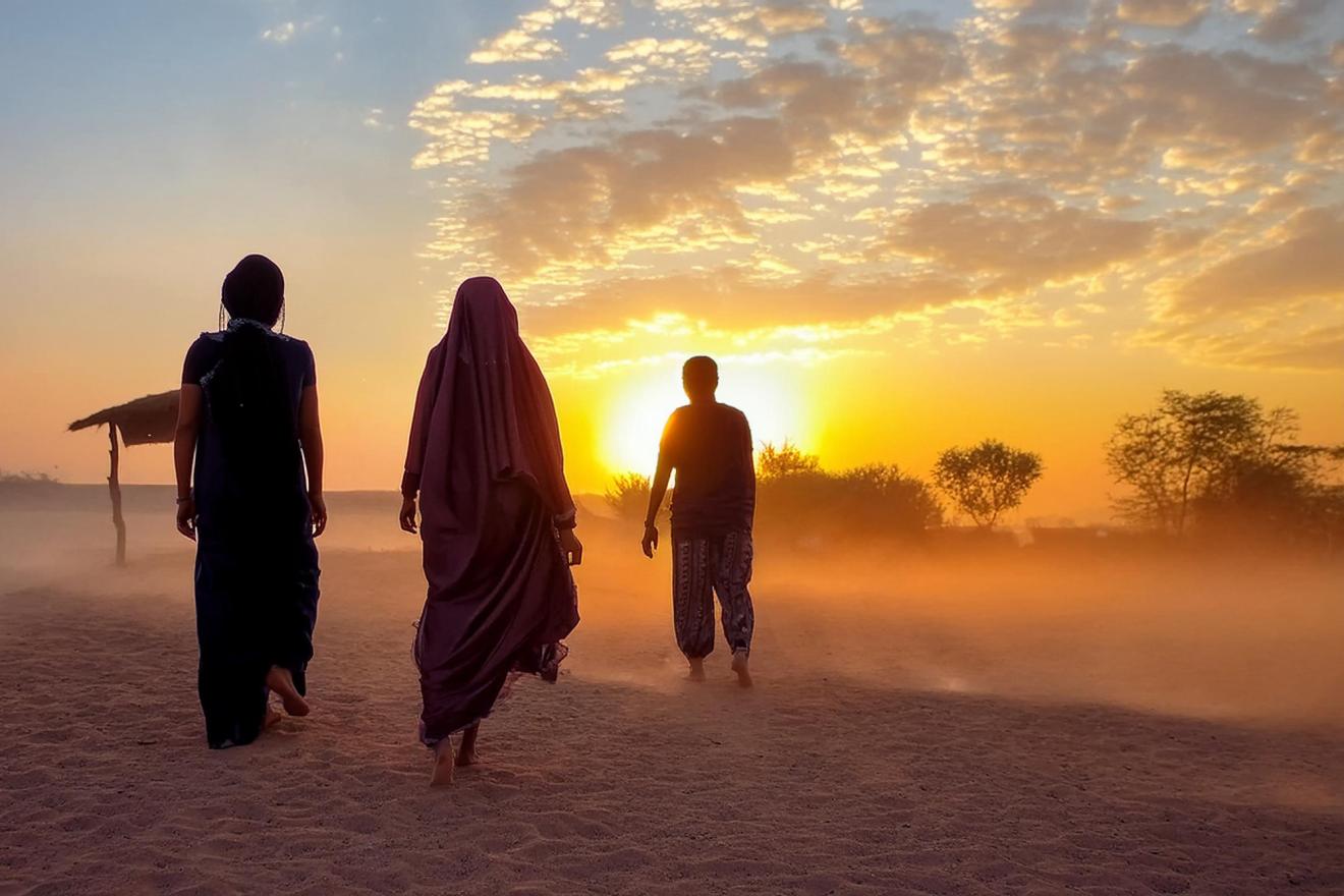 Three people walk across the desert, the sun hanging on the horizon ahead.