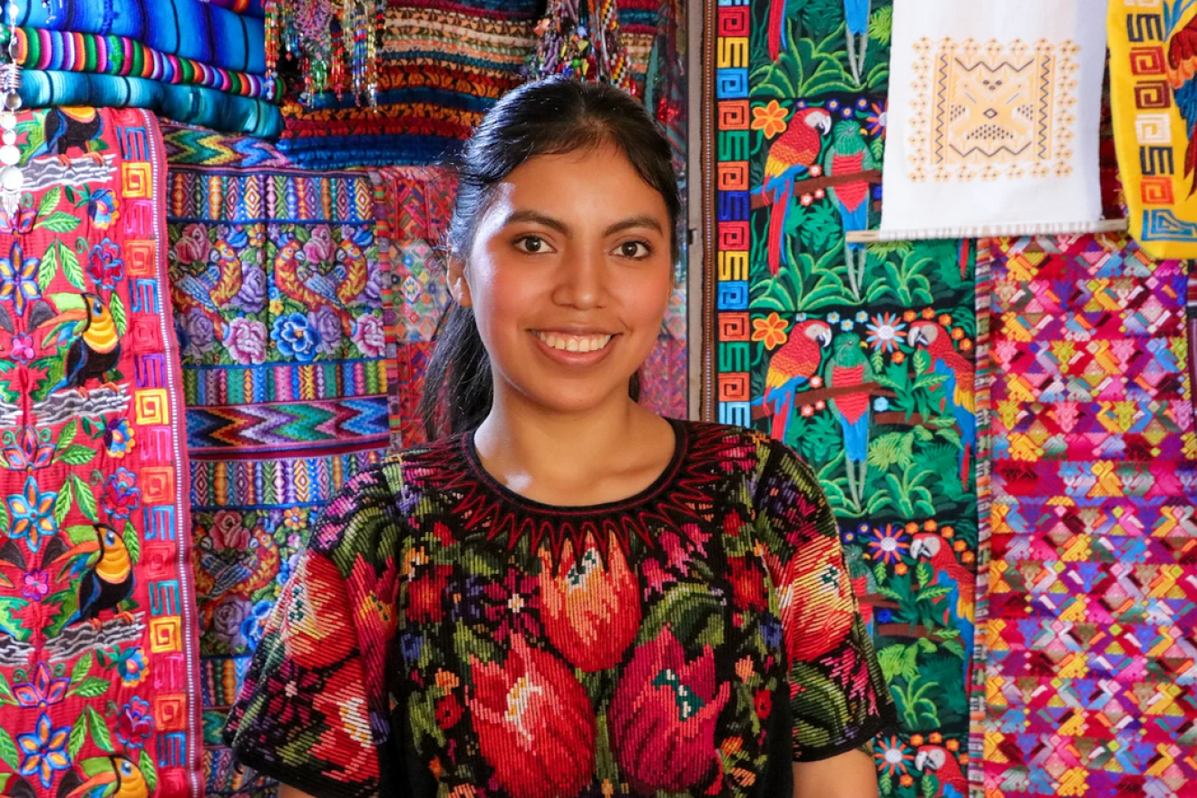 Una joven sonríe frente a vestidos tradicionales de su creación. 
