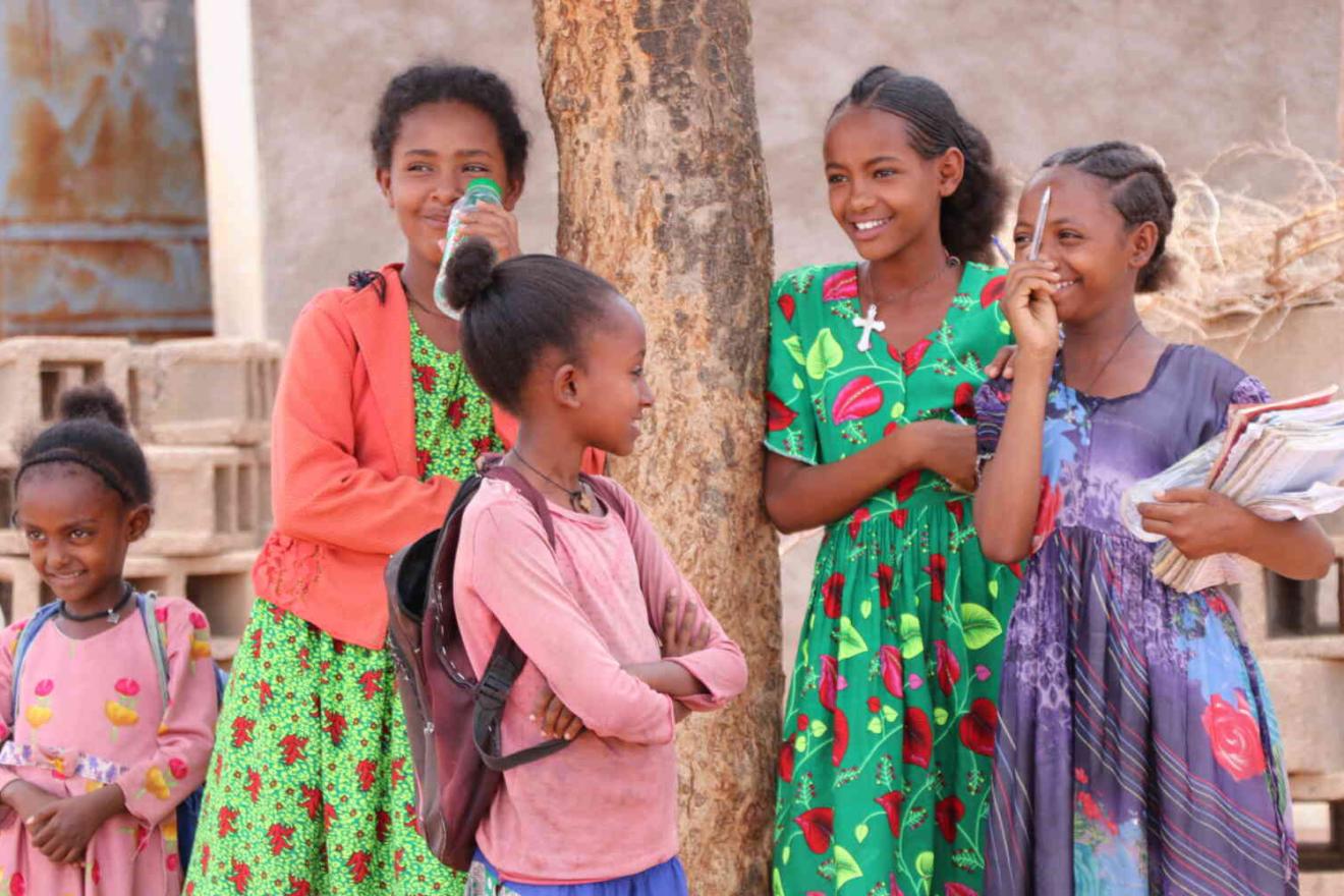 Un groupe d'enfants debout à l'extérieur près d'un arbre, portant des vêtements colorés et tenant des livres et des sacs à dos