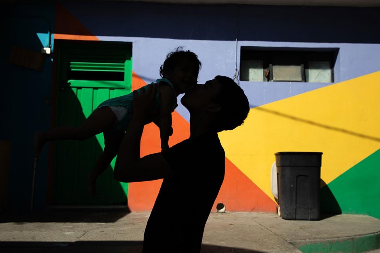 Un homme tient entre ses bras un enfant, en contre-jour, devant un mur coloré