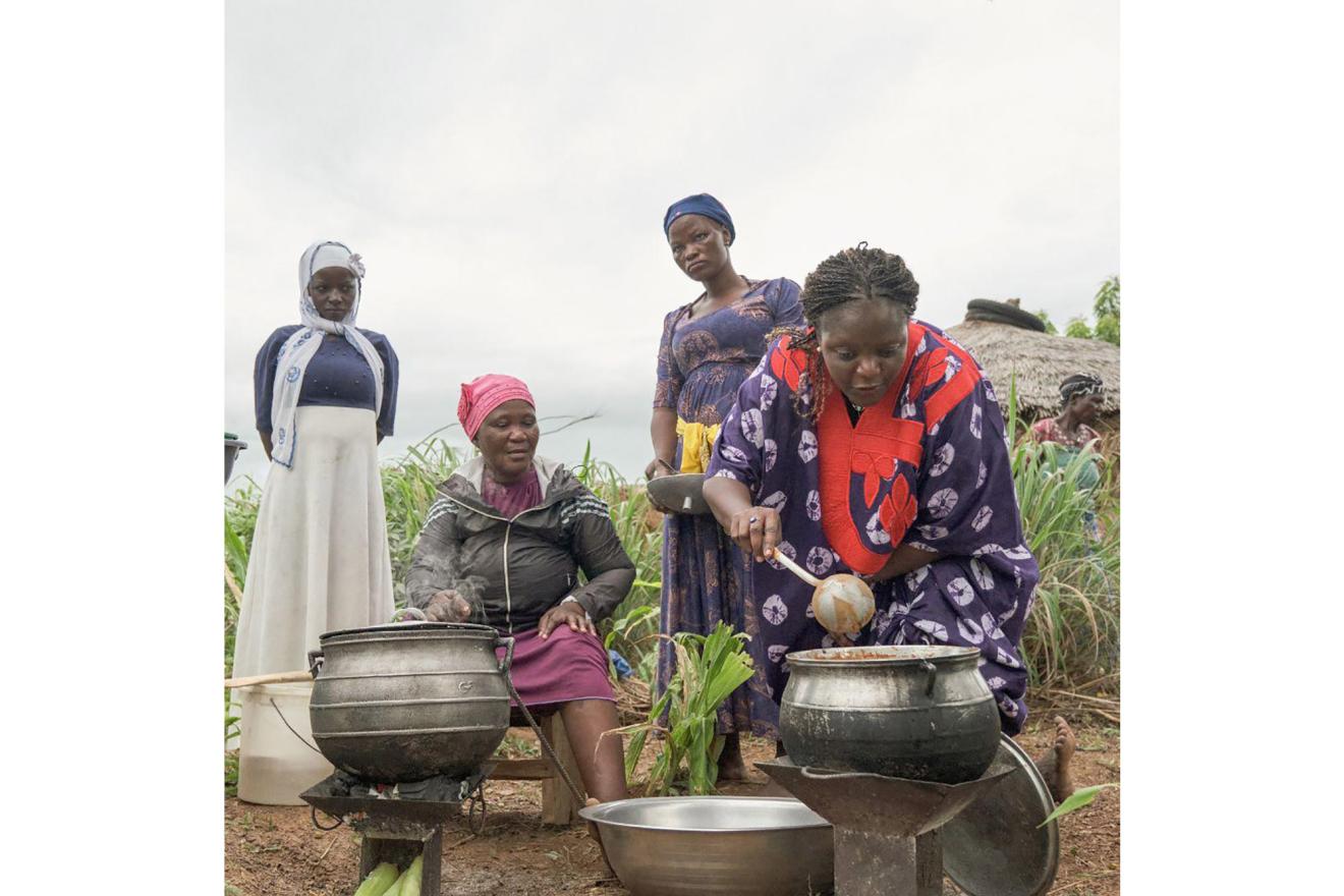 Four women cooking outdoors, with three standing and one sitting. The person sitting is stirring a pot on a stove, while another person is also attending to a pot.