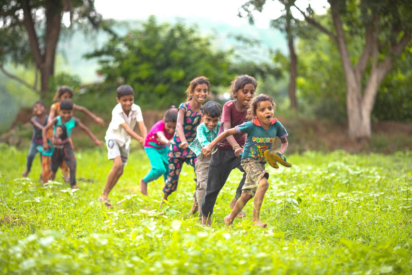 Children run joyfully through a lush green field, holding hands.