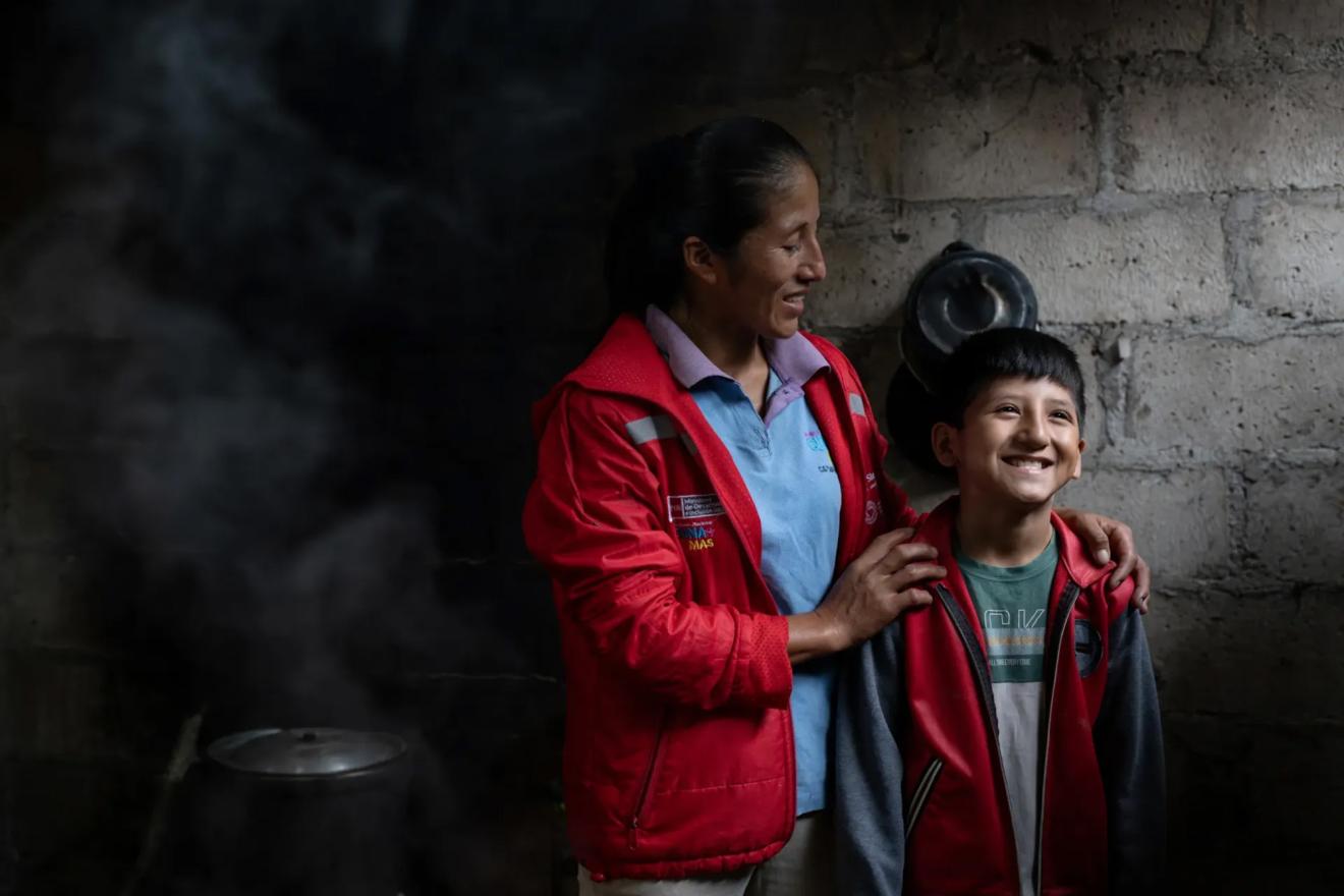 A woman gently placing her arm on the shoulder of a child in a dimly lit, rustic setting.