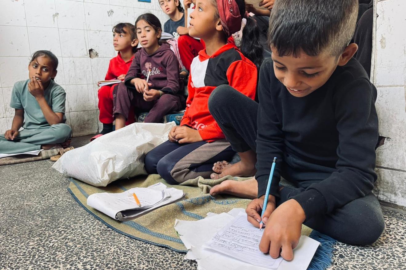 Un groupe d'enfants sont assis serrés les uns contre les autres sur le sol usé d'une petite pièce carrelée, écrivant et écoutant attentivement pendant qu'ils étudient malgré le cadre rudimentaire.