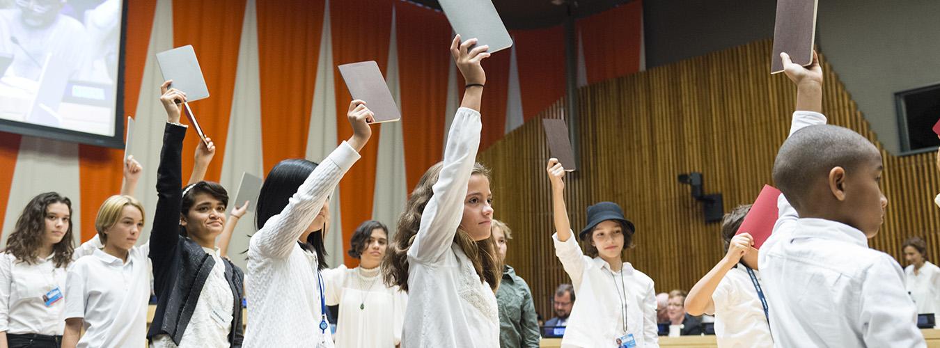 A group of children in a UN Hall stand up holding up notebooks. 