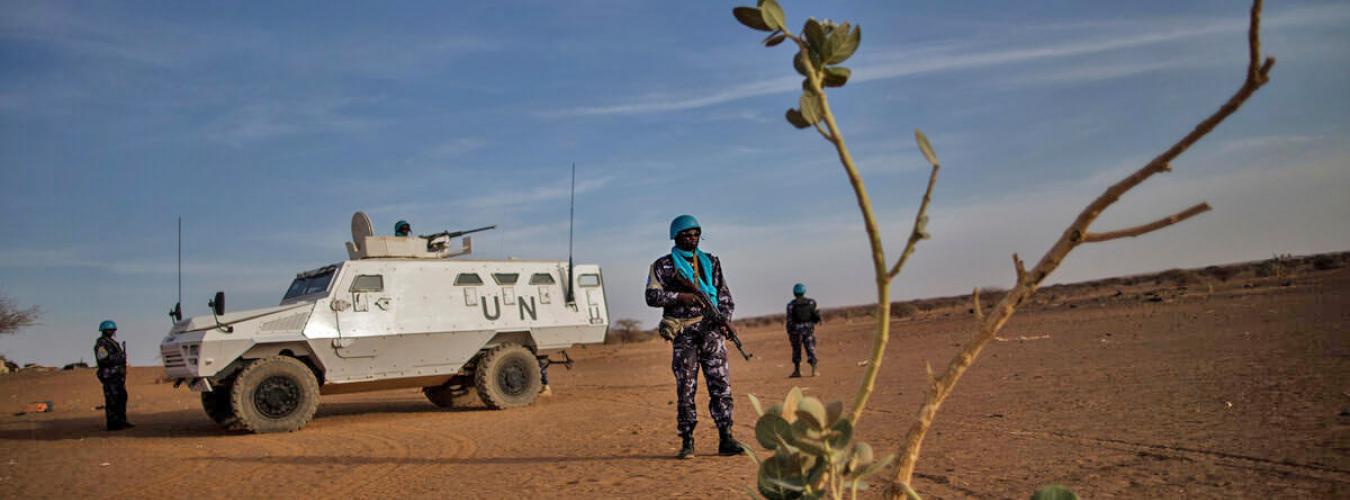 Un miembro del personal de mantenimiento de la paz de la ONU haciendo guardia con 2 miembros del personal de mantenimiento de la paz y un vehículo de la ONU al fondo.