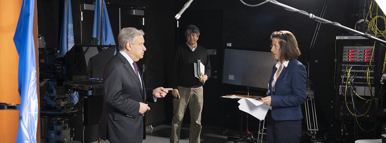 UN Secretary-General António Guterres stands in TV studio with Melissa Fleming holding a script and a technician in the background.