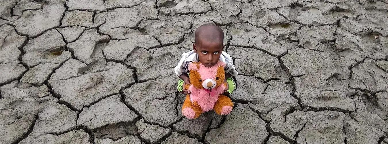 A little boy with his teddy on a soil affected by drought.