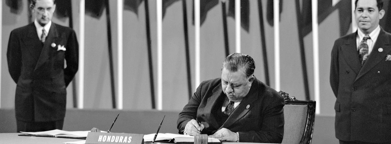 El presidente de la delegación de Honduras, sentado en primer plano, firmando la Carta de la ONU en 1945 con dos miembros de la delegación al fondo, mirando.