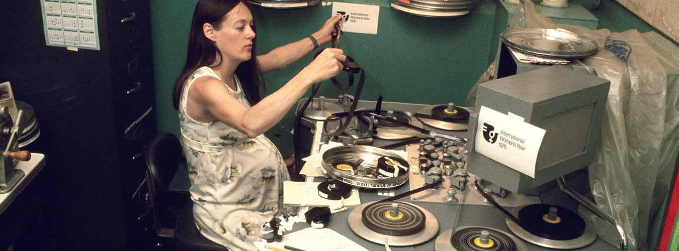 Pregnant woman look at film reels. The symbol for international Women's Year can be seen in the background and on the speaker to the right.