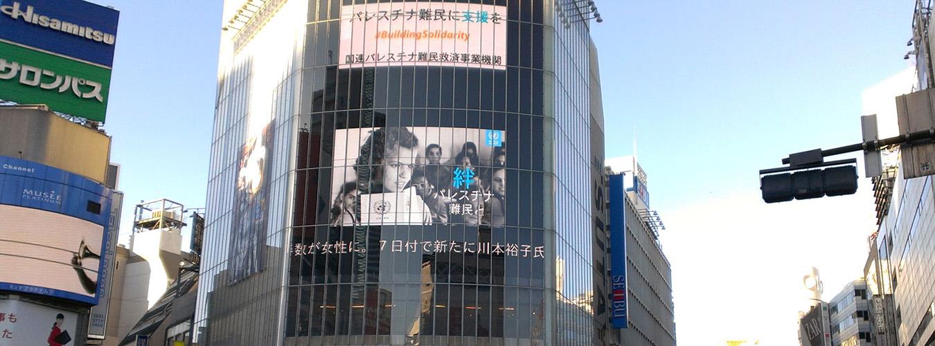 Valla publicitaria electrónica en el centro de Tokio con una imagen de la UNRWA y el lema #ConstruyendoSolidaridad
