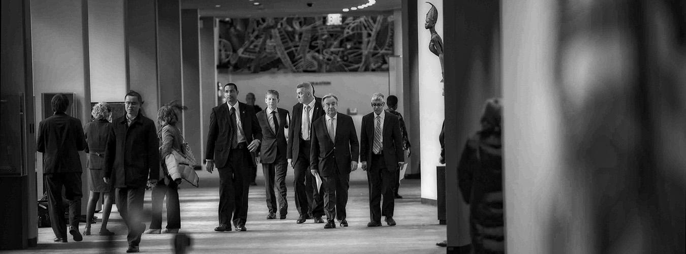Black and white image of a group of men walking through a hall in UN headquarters in New York.