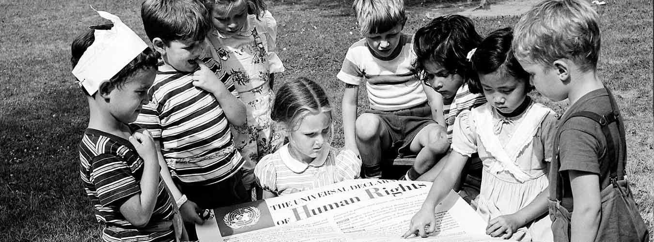 Archival balck and white photo of children standing around a large poster of the UDHR.