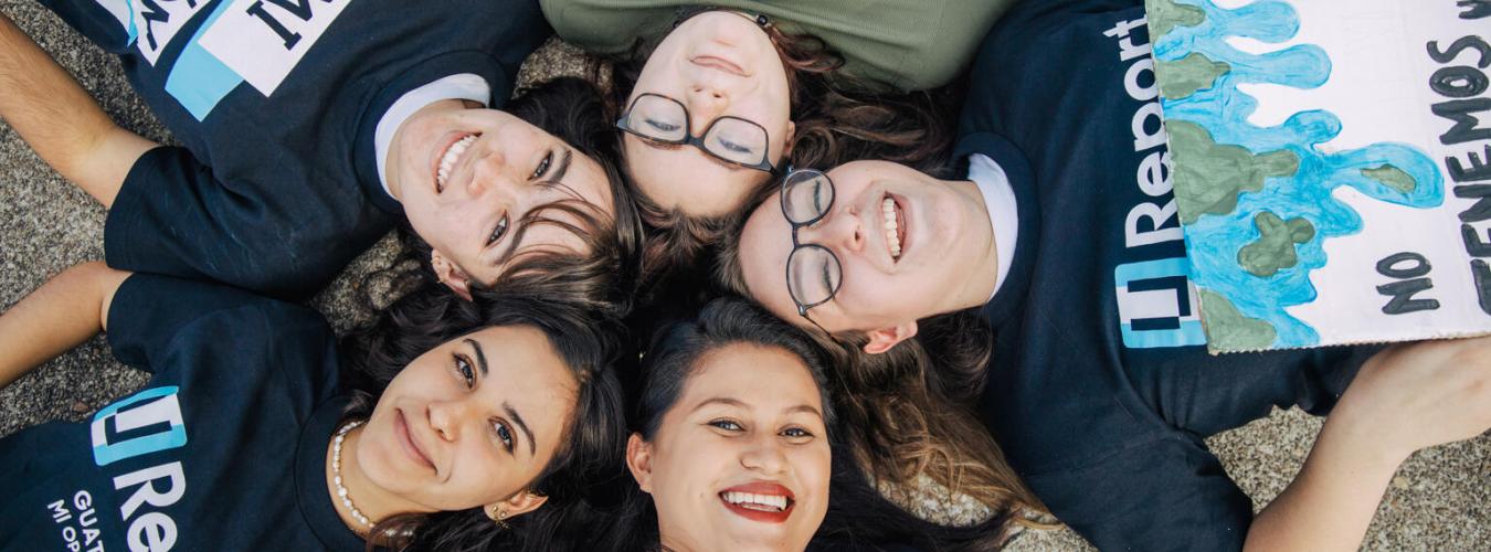 Five young women working on a project lay on their backs smiling up at the camera
