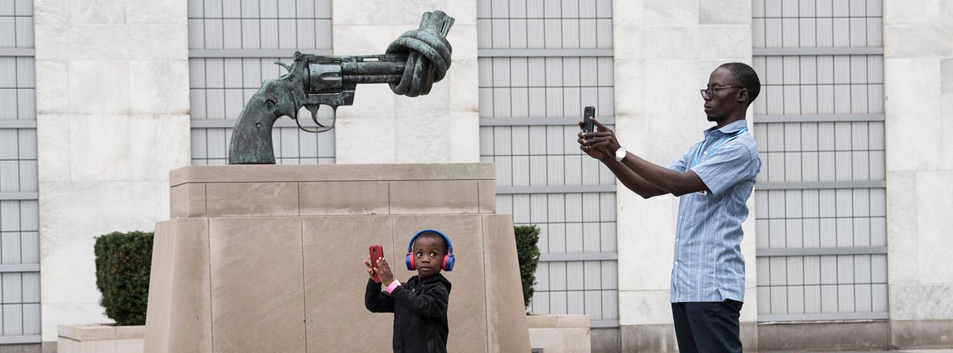Un adulto y un niño sosteniendo teléfonos móviles y haciendo una foto junto a una escultura de un arma con el cañón anudado, en la Sede de la ONU en Nueva York.