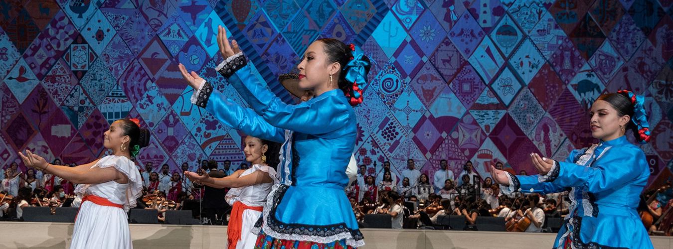 Una bailarinas en un escenario, llevan coloridos vestidos.  