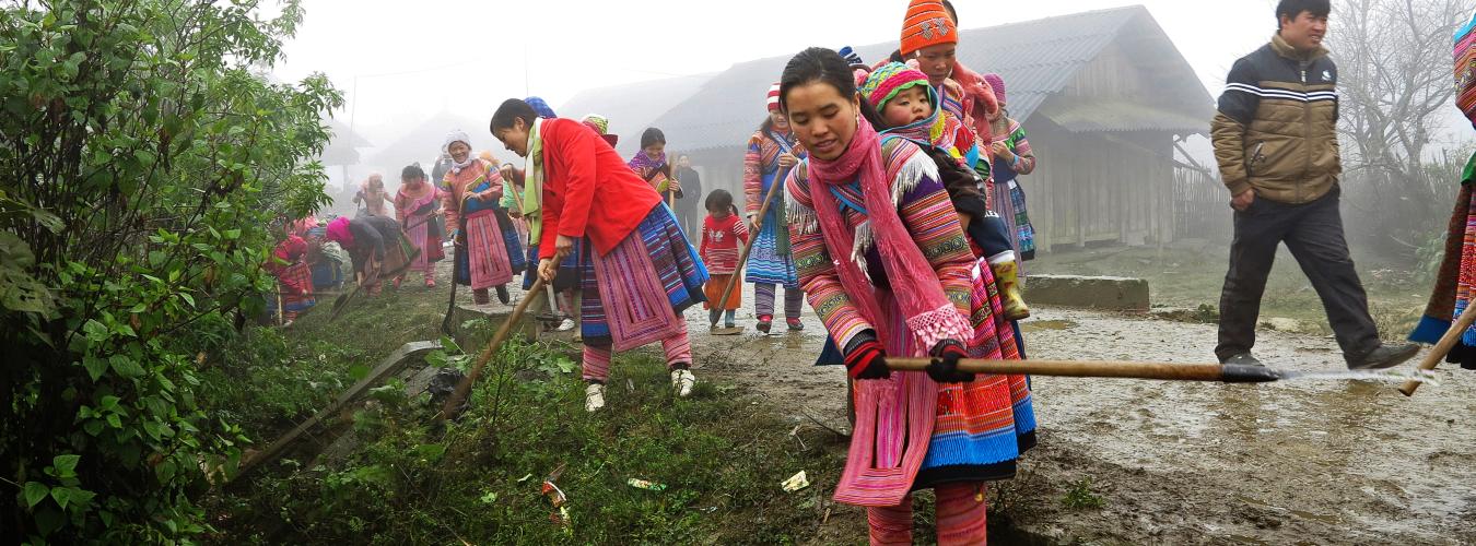 women in traditional outfits clear debris from the side of the road with a sickle.