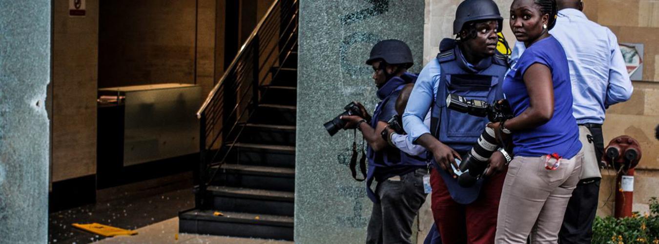 Periodistas con equipo de protección frente a un edificio con la entrada de cristal rota.