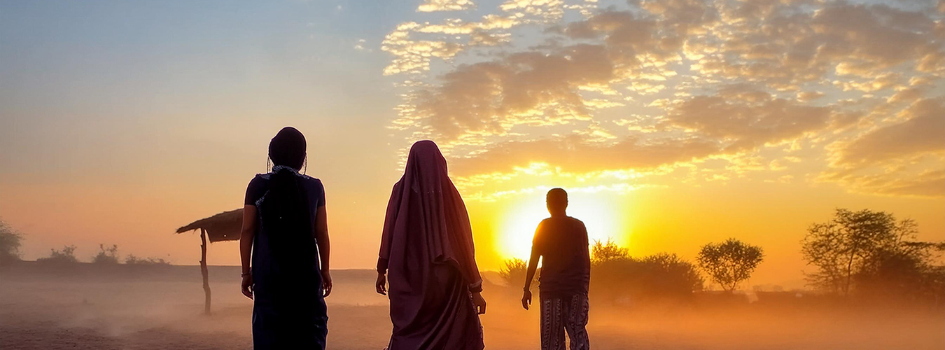 Three people walk across the desert, the sun hanging on the horizon ahead.