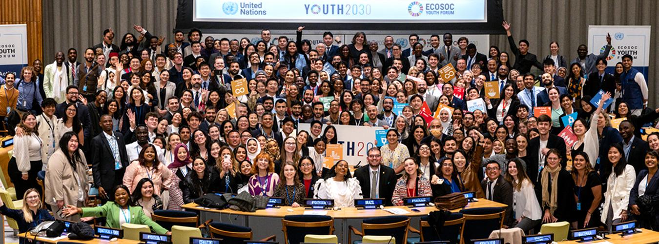 a large group of youth in the UN ECOSOC chamber raise their hands for a group photo
