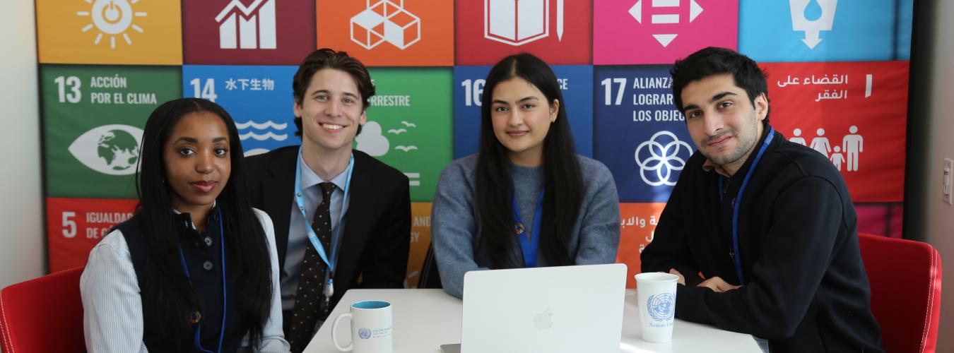 Four UN interns sitting around a white table with a laptop and UN-branded mugs, in front of a colorful backdrop displaying the United Nations Sustainable Development Goals.