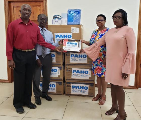 Two women and two men stand facing the camera as they flank stacked up boxes with the label "PAHO".