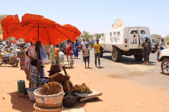 In a busy street produce vendors shelter under a large red umbrella as UN Blue Helmet police stand guard.