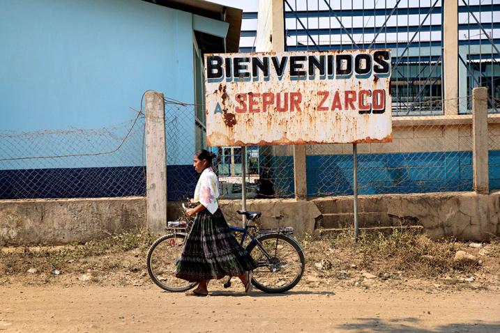 A woman walking with a bike in front of a board that says Welcome to Sepur Zarco
