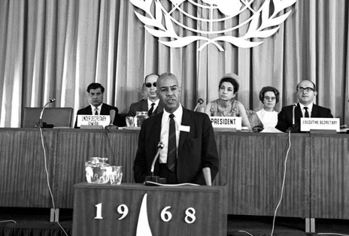 Roy Wilkins speaking on the podium with participants seated at a table behind him.