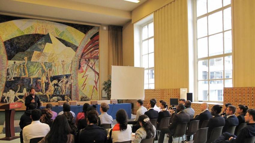 People are seated at the carpeted open space of the library with a large wall mural. 