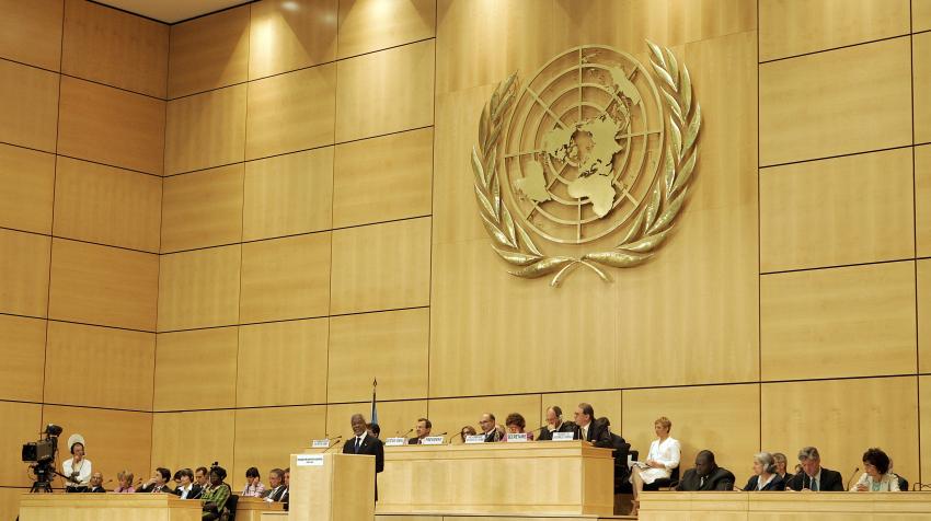 Frontal view of the Assembly Hall with UN emblem on bright colored wooden wall and a set of podiums and tables in the front. 