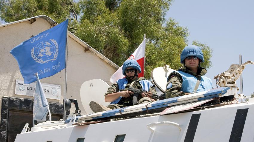 Two Polish peacekeepers are riding on a white patrol vehicle with the Polish national flag and the UN flag.