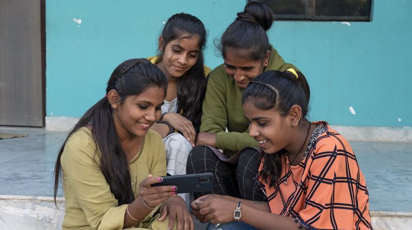 Group of adolescent girls seated on steps look at mobile phone screen.