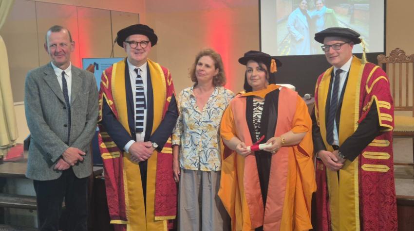 Five people at a ceremony, including Atefa Waseq in academic robes holding a diploma, flanked by two university officials in red and gold gowns.