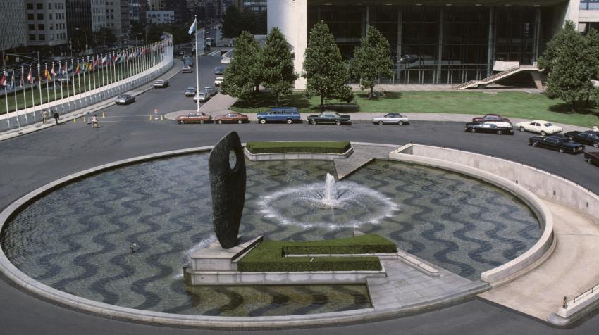 Overview of a circular pool with a fountain in the center, formed of alternating bands of crushed white marble and black pebbles, and an abstract sculpture at the edge of the pool. 