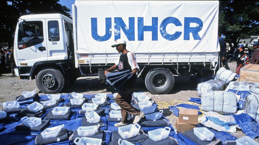 A UNHCR worker is preparing repatriation kits, including plastic sheeting, a blanket, a gerrycan, and soap, in front of a truck with the UNHCR logo on it. 