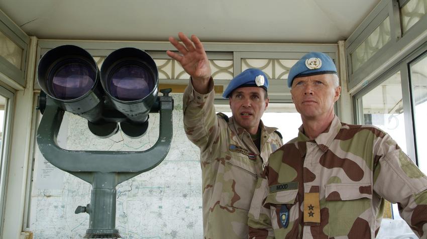 Two male soldiers from the peacekeeping mission are standing next to binoculars at an observation post. 