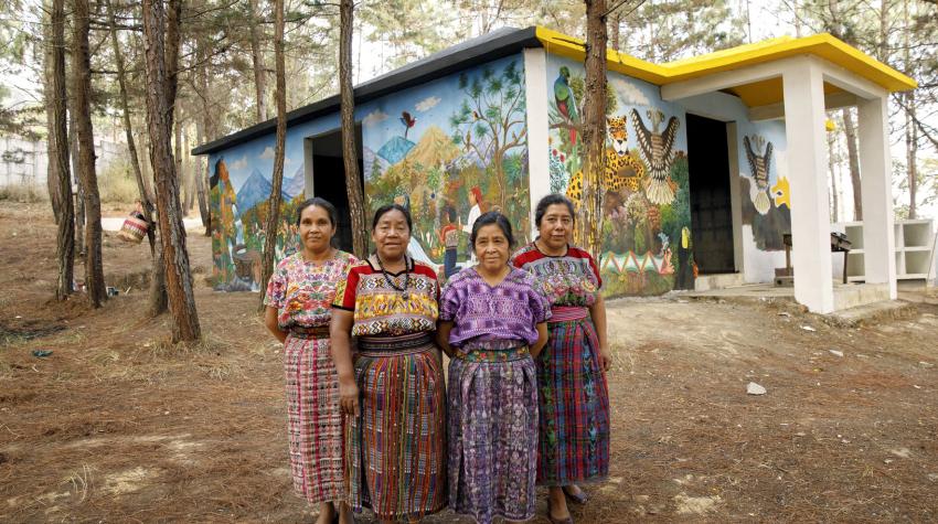 4 women artists stand in front of a hand-painted memorial for female victims of the conflict in Comalapa, Guatemala