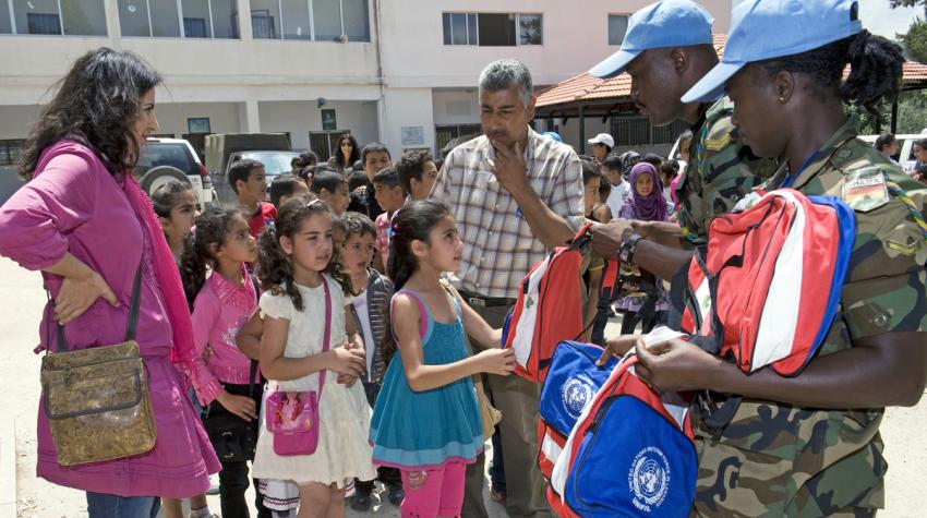 Two peacekeepers are delivering school backpacks to school children lined up. 