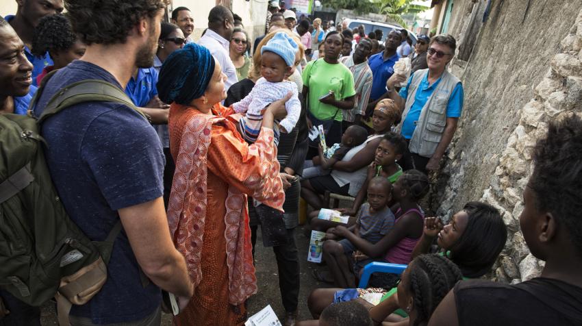 Woman, surrounded by crowd of people, is holding a baby in her arms.