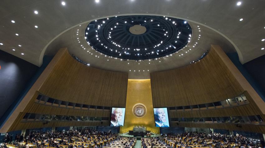 Bottom to top view of the General Assembly Hall with its round paved ceiling, golden part of the wooden wall with the UN emblem, and several rows of seats.