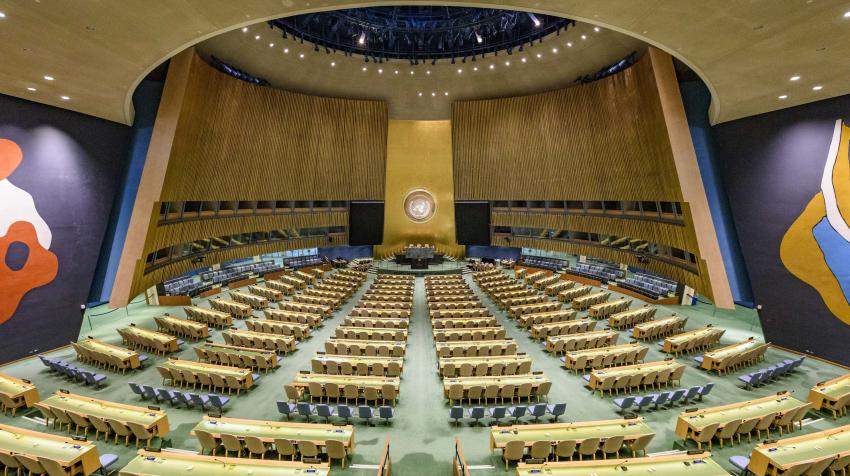 Overview of the General Assembly Hall, showing several rows of tables and seats, and murals on both sides of the back wall. 