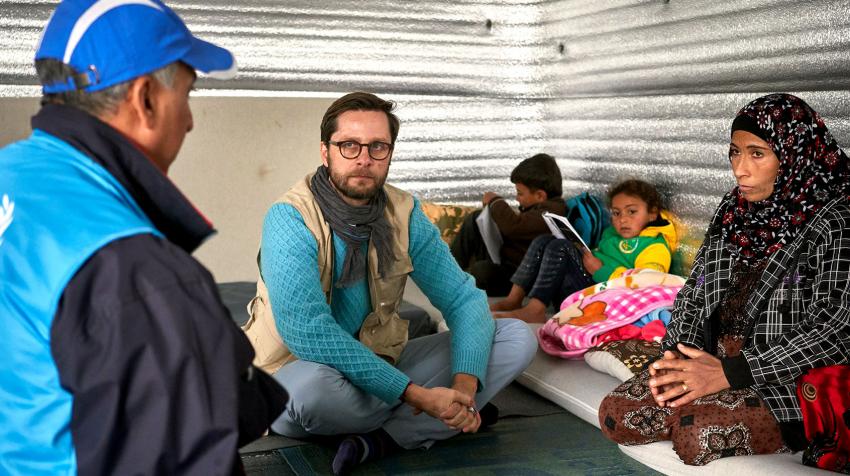 Three adults and two children are sitting on the floor in a refugee makeshift home. The adults are talking and look concerned.