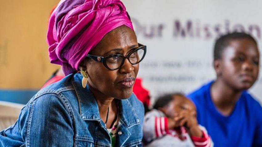 Winnie is wearing a beautiful pink head-wrap and is seated, leaning forward, as she listens intently with children seen in the background.