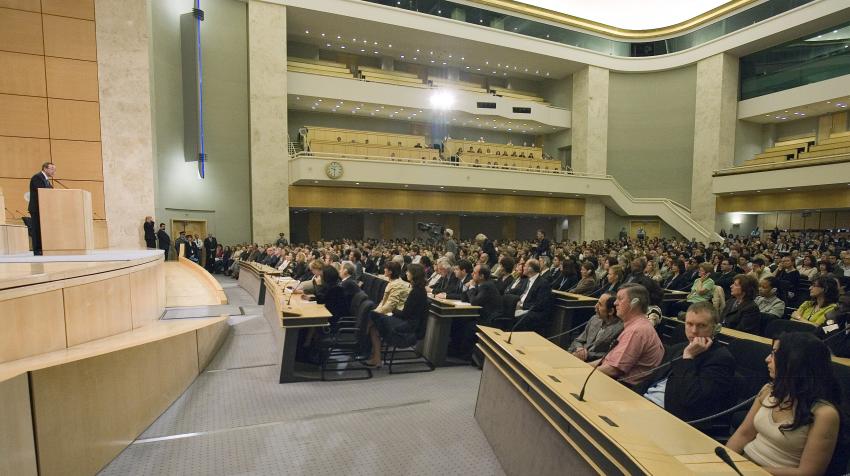 Floor level view of man speaking at podium and audience listening below, behind having 2 more floors for seats.  