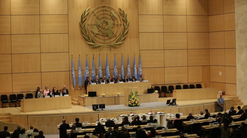 Front view of the Assembly Hall with several rows of seats and a stage with elevated tables and series of UN flags under UN emblem. 