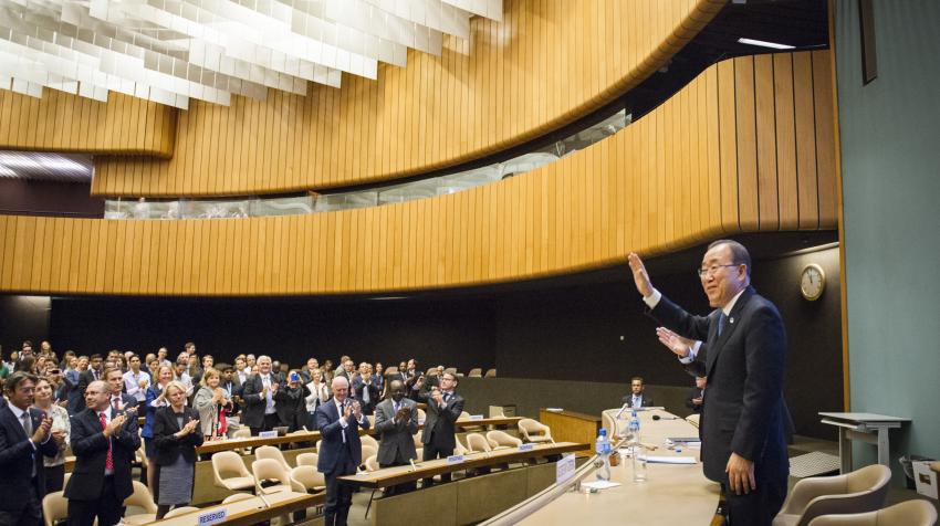 Former Secretary-General Ban Ki-moon waving in front of the Hall while a standing audience applauds. The Hall's walls are partially composed of wood with a ceiling composed of rows of hanging white tiles. 