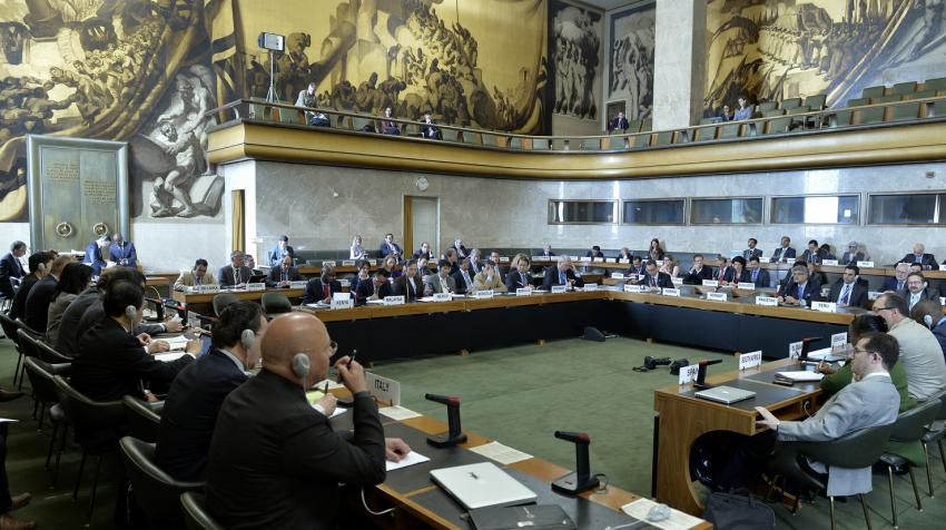 Individuals sit in square green-carpeted room with row of desks facing center