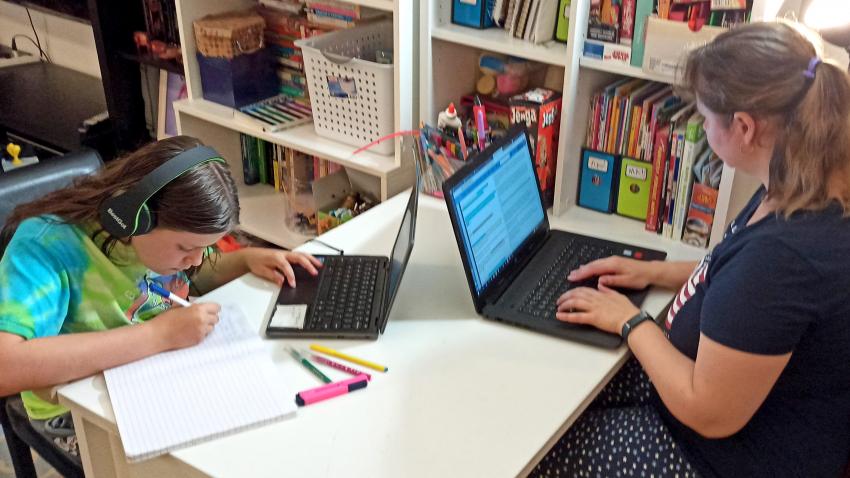 Woman and a girl share a desk while working on their laptops.