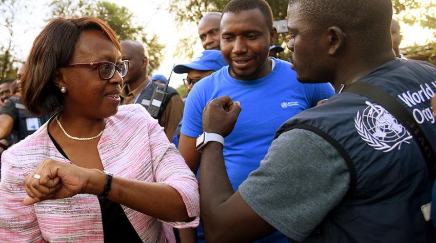 Dr. Moeti does an elbow shake with a man wearing a vest that says "WHO"